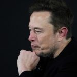 Elon Musk on stage at an event, resting his chin on his hand