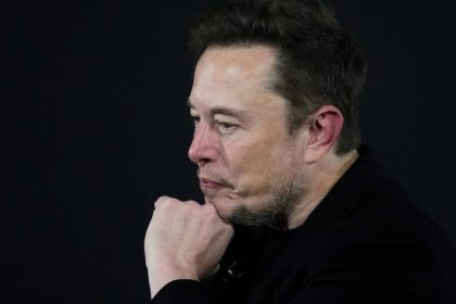 Elon Musk on stage at an event, resting his chin on his hand