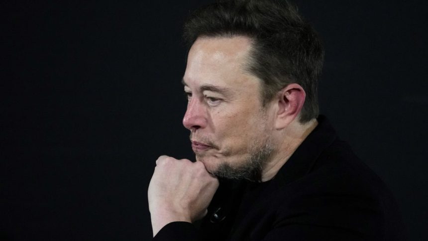Elon Musk on stage at an event, resting his chin on his hand