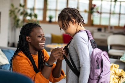 A smiling mother adjusts backpack straps for a young child in a living room.